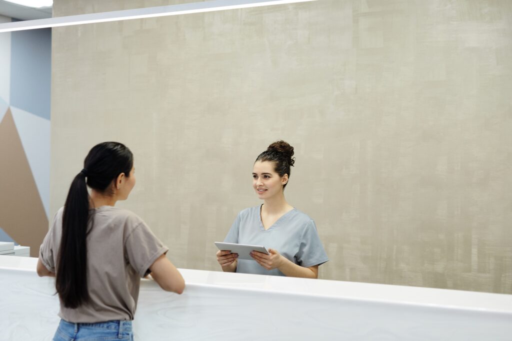 Medical receptionist using a tablet while assisting a patient at the front desk of a healthcare clinic.
